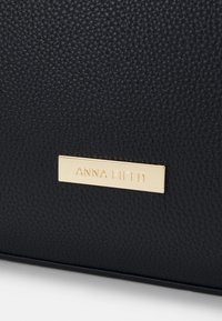 Surface en cuir noir texturé avec une petite plaque métallique rectangulaire dorée gravée du nom de la marque "ANNA FIELD."