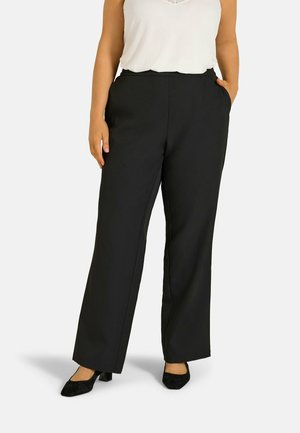 Femme portant un pantalon noir à jambes larges, un haut blanc sans manches et des chaussures noires à talons bas, debout avec une main dans la poche.