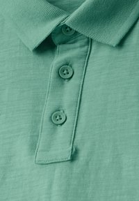 Camisa polo verde menta con una tela texturizada, que presenta una tapeta con botones y tres botones a juego. Cuello plegado y detalles cosidos.