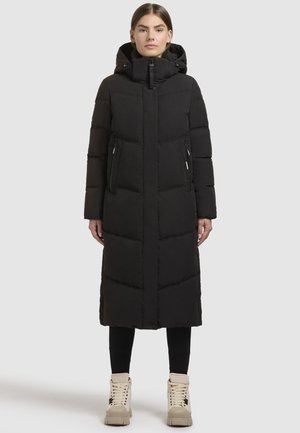 Cappotto lungo nero imbottito con cappuccio, caratterizzato da una chiusura con zip, tasche laterali con zip e motivo a righe orizzontali. Indossato sopra leggings neri e stivali beige.