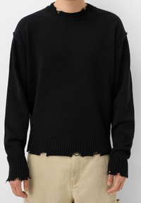 Svart stickad tröja med ribbad halsringning och nederkant, med slitna kanter vid ärmsluten och nederkanten. Standard passform med långa ärmar.