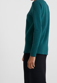 Türkisfarbenes langärmliges Shirt mit rundem Halsausschnitt, aus weicher Baumwolle, mit geradem Saum und glatter Textur, seitlich dargestellt.