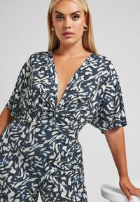 Keksikuvioinen haalari tummansinisessä ja kreemassa, syvällä V-pääntiellä, lyhyillä hihoilla ja kääntöyksityiskohdalla, valmistettu kevyestä kankaasta.