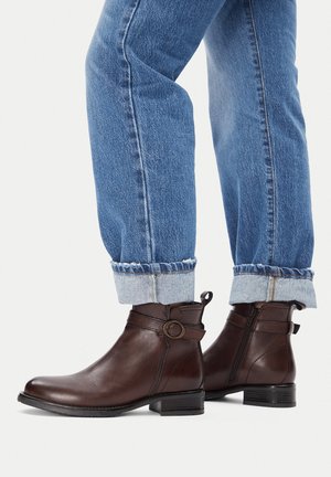 Smeđe kožne gležnjače sa zaobljenim vrhom, bočnim patentnim zatvaračem i ukrasnim remenom s metalnim detaljem u obliku kruga. Nošene uz plave traperice s preklopom.