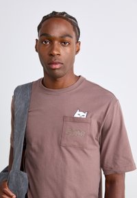 Tricou din bumbac maro, cu un buzunar pe piept decorat cu un design al unei pisici albe și logo-ul "PUMA" în font bej deschis. Mâneci scurte.