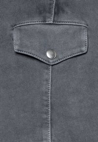 Tela de mezclilla gris con un bolsillo puntiagudo con cierre de botón metálico, costuras visibles y una superficie lisa con una textura ligera.