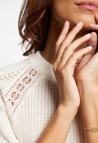 Frau in cremefarbener Strickjacke mit Spitzenakzenten, die mit der Hand ihr Kinn berührt und ein zartes goldfarbenes Armband am Handgelenk trägt.