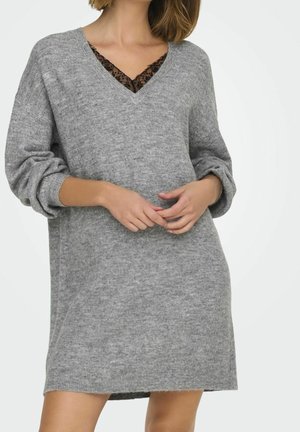 Robe-sweater oversize grise en tricot avec un décolleté en V, présentant une bordure en dentelle noire visible en dessous. Manches longues à revers.