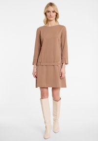 Braunes, langärmeliges Kleid mit Rundhalsausschnitt, taillierter Taille und seitlichen Schlitzen; glattes Material und minimalistisches Design. Kombiniert mit beigefarbenen Stiefeln bis zum Knie.
