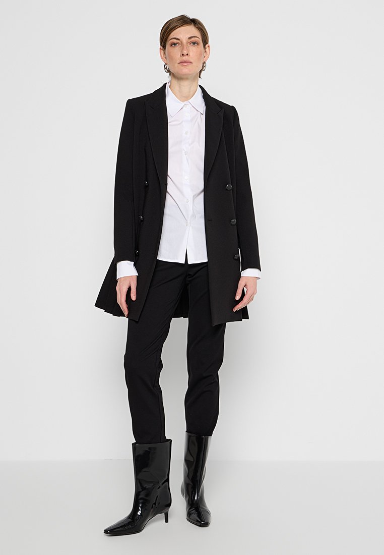 Schwarzer Überwurf mit doppelt-knöpfbaren Knöpfen, getragen über einem weißen Hemd und schwarzen Hosen. Glänzende schwarze kniehohe Stiefel vervollständigen das Outfit.