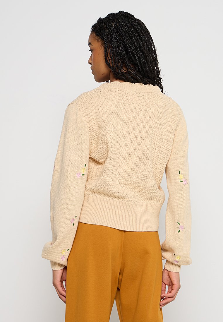 Crème gebreide trui met een gestructureerd patroon, geribbelde manchetten en geborduurde bloemaccenten op de mouwen, gecombineerd met mosterdkleurige broek.