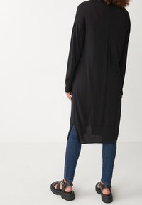 Długi czarny dzianinowy kardigan z bocznymi rozcięciami, ściągaczami na rękawach i luźnym krojem, zestawiony z ciemnoniebieskimi dżinsami i czarnymi sandałami na platformie.