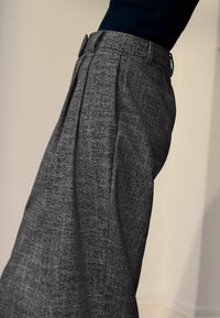 Persona che indossa pantaloni grigi a vita alta con texture e pieghe frontali, abbinati a una maglietta nera, mostrata di lato su uno sfondo semplice.