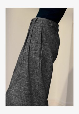 Asmuo, vilkintis aukšto juosmens pilkas tekstūruotas kelnės su priekiniais raukiniais ir juoda palaidine, pavaizduotas iš šono prieš paprastą fono.