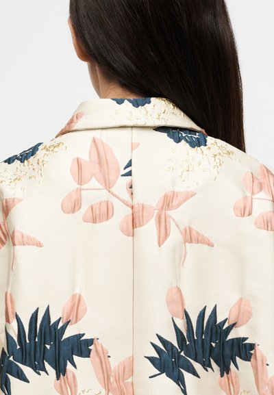 Veste à motif floral crème, ornée de feuilles roses et bleu marine, avec une texture lisse et un subtil accent doré le long du col.