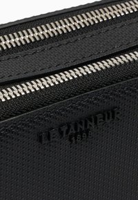Pochette en cuir texturé noir avec deux zippers en argent et le logo embossé "LE TANNEUR 1898". Présente un motif géométrique raffiné.
