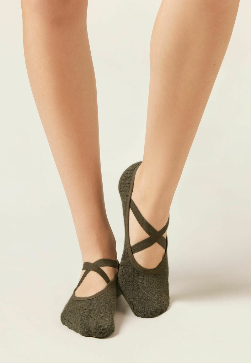 Jambes portant des chaussettes à doigts style ballet vert foncé avec des brides croisées sur un fond clair, pieds posés avec grâce.