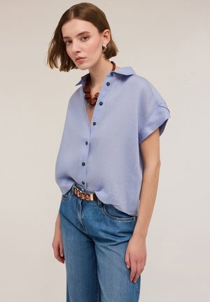 Camicia azzurra a maniche corte con colletto, con chiusura a bottone caratterizzata da bottoni neri, abbinata a jeans blu a vita alta.