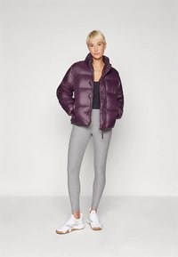 Lila vadderad jacka med glänsande yta, utrustad med hög krage och knappar fram, kombinerad med grå leggings och vita sportskor.