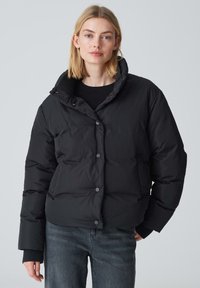 Giacca a vento nera con colletto alto, dotata di bottoni a pressione e tasche laterali. Il materiale è liscio e imbottito per l'isolamento termico.