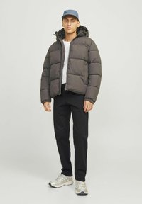 Dunkelgraue Daunenjacke mit horizontalem Steppmuster, Reißverschluss und Kapuze. Kombiniert mit schwarzen Hosen und hellgrauen Sportschuhen.
