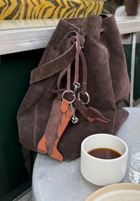 Brązowa torba typu bucket zamszowa z brelokami oraz filiżanka czarnej kawy na metalowym stole obok poduszki w tygrysi wzór.