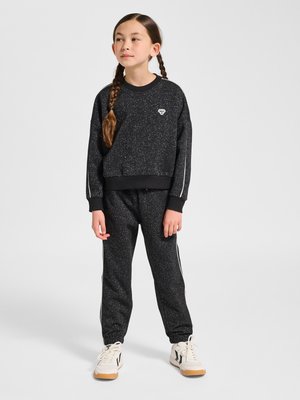 Młoda dziewczyna z warkoczami, ubrana w czarną, cętkowaną bluzę oraz pasujące dresowe spodnie z białymi paskami i białe buty sportowe.