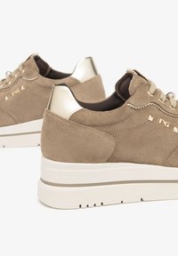 Baskets en suédine beige avec de épaisses semelles blanches, embouts de talons dorés métalliques et accents de clous dorés près des lacets sur fond blanc uni.