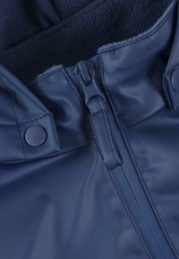 Navy blue vesipitävä takki, jossa on vetoketjukiinnitys, pehmeä fleecevuori ja napinläpipainikkeet varmassa kiinnittämisessä.