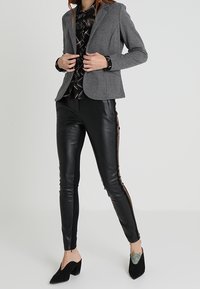 Blazer grigio in tessuto morbido, blusa nera a fantasia, pantaloni in pelle nera con dettagli in cerniera laterale e décolleté neri.