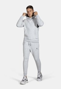 Felpa grigia con cappuccio e pantaloni jogger coordinati in tessuto morbido. Presenta una tasca frontale, polsi a costine e dettagli di logo neri. Indossata con sneakers.