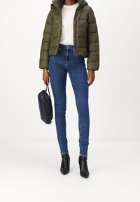 Olijfgroene pufferjas met capuchon, een wit t-shirt, donkerblauwe skinny jeans en zwarte enkellaarzen. Een marineblauwe tas vasthoudend.