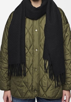 Personne portant une veste matelassée vert olive avec des boutons-pression et une épaisse écharpe noire texturée aux extrémités frangées.