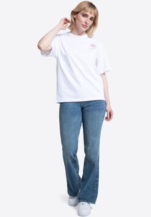 Mujer con camiseta blanca con diseño gráfico, vaqueros azules y zapatillas blancas, de pie con una mano cerca de la cabeza sobre un fondo blanco.