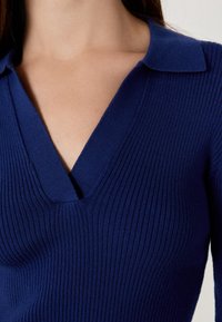 Zbliżenie osoby w prążkowanym, ciemnoniebieskim swetrze w serek z kołnierzykiem, ukazujące dekolt i górną część tułowia.