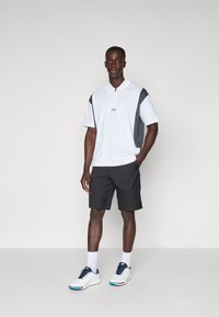 Hvit polo med kort erme og grå sidepaneler; svarte shorts; hvite sneakers med marineblå detaljer. Modell står foran en ensfarget bakgrunn.