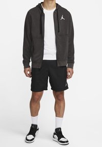 Felpa con zip grigia scura con logo, T-shirt bianca, pantaloni corti neri e sneakers bianco e nere. Presenta un design atletico casual.