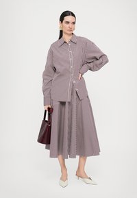 Camicia a righe bordeaux e bianche, vestibilità oversize, design con bottoni. Abbinata a una gonna a righe coordinata e una borsa bordeaux. Tacchi bianchi.