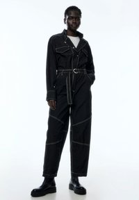 Schwarzer Jumpsuit mit kontrastierendem weißen Nähten, tailliertem Gürtel und großen Fronttaschen. Hergestellt aus glattem Stoff mit strukturiertem Schnitt.