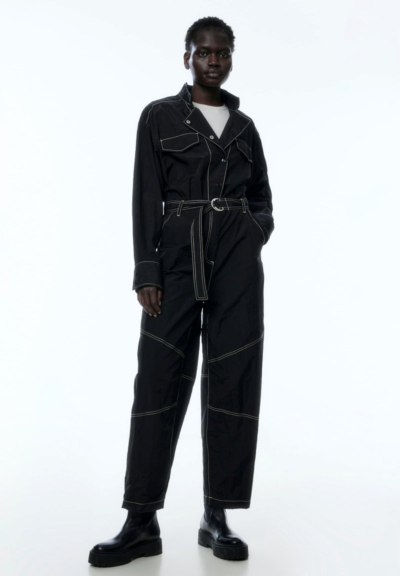 Schwarzer Jumpsuit mit kontrastierendem weißen Nähten, tailliertem Gürtel und großen Fronttaschen. Hergestellt aus glattem Stoff mit strukturiertem Schnitt.