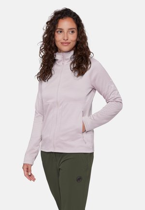 Kvinde med krøllet hår iført en lyse lilla lynlåstrøje og olivengrønne bukser, stående med den ene hånd i lommen og kigger til siden.