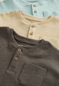 Tres camisetas Henley en marrón, beige y azul claro. Cada una presenta una tela de punto texturizada, tapeta de botones y un detalle de bolsillo en el pecho.
