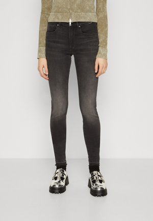 Persoon draagt versleten zwarte skinny jeans, een beige jas met ritssluiting, zwarte sokken en zilveren schoenen met chunky hak en gespen.