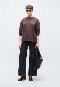 Barna pulóver "DOLCE FAR NIENTE" felirattal, sötét, bővülő fazonú farmerrel és fekete szövött táskával párosítva. Simaságú anyag és laza szabás.