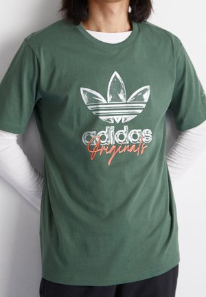 Grøn bomulds-t-shirt med stort hvidt Adidas trefoil-logo og rød tekst "Originals". Kortærmet med rund hals.