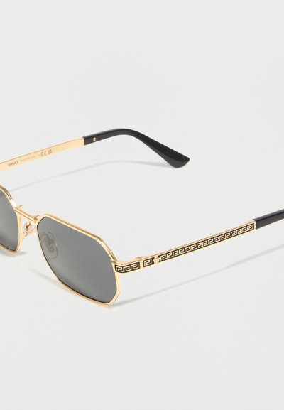 Γυαλιά ηλίου με χρυσή κορνίζα, μαύρους βραχίονες με γεωμετρικό μοτίβο και σκουρόχρωμους εξάγωνους φακούς σε λευκό φόντο.