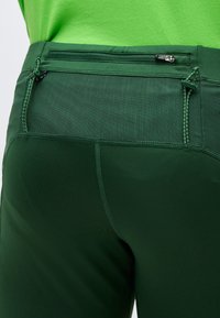 Κοντινό πλάνο σε πράσινο αθλητικό παντελόνι με δίχτυ στο πίσω μέρος και ασφαλή τσεπάκι με φερμουάρ στο κάτω μέρος της πλάτης, φορεμένο με φωτεινή πράσινη μπλούζα.