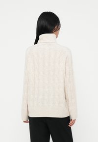 Kvinne med langt svart hår iført en off-white kabelstrikket høyhalset genser og svarte bukser, vendt bort fra kameraet mot en hvit bakgrunn.