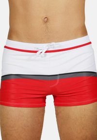Un short de bain rouge et blanc avec une bande grise horizontale, fabriqué à partir d'un matériau extensible avec une taille à cordon de serrage et des coutures contrastées.