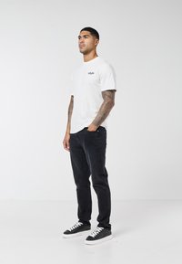 Mand med tatoverede arme iført hvid grafisk t-shirt, sorte jeans og sorte sneakers, står op ad en ensfarvet hvid baggrund.